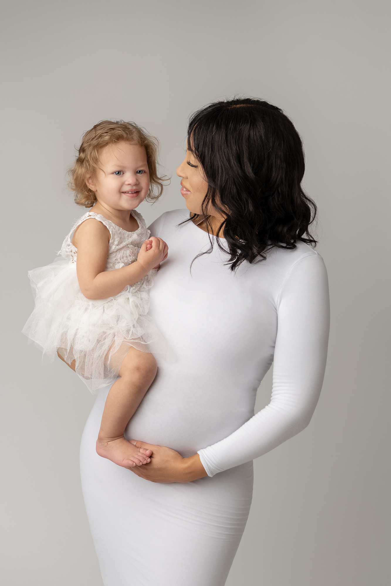 mom to be holding her daughter on her hip both dressed in white