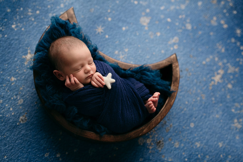 newborn baby boy wrapped in blue holding a star in a moon shaped bed