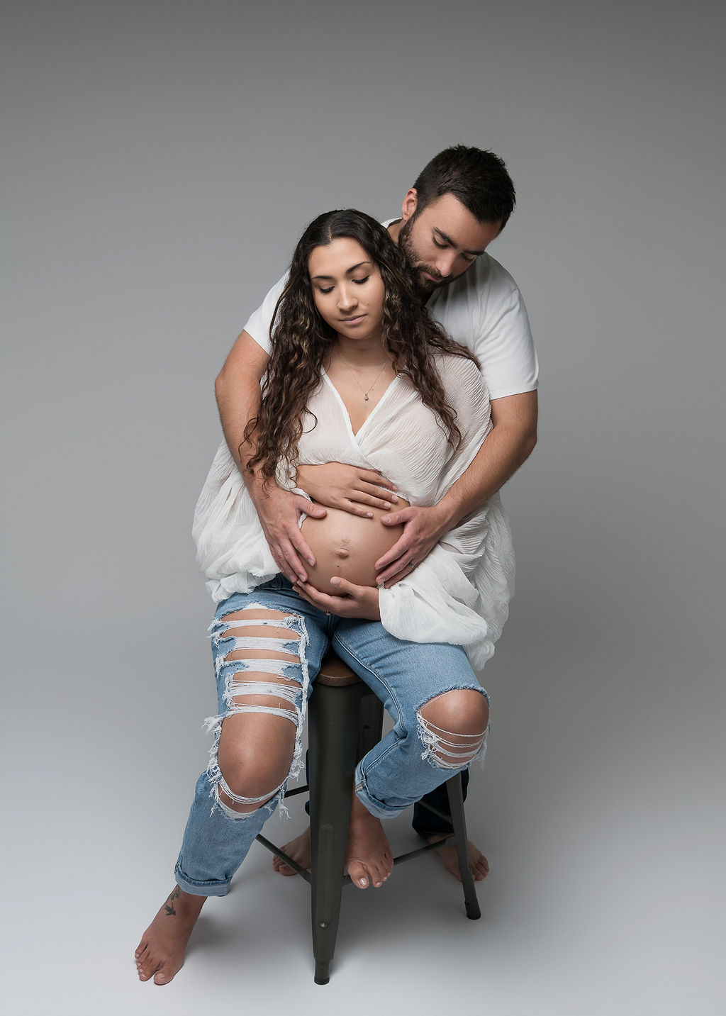 mom to be sitting on a stool with the father's hands on her belly